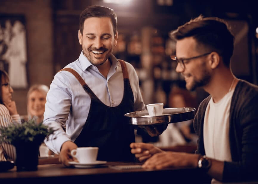Serveur souriant servant un café à un client dans un café chaleureux – image représentant l’accueil et la convivialité dans un établissement HCR.